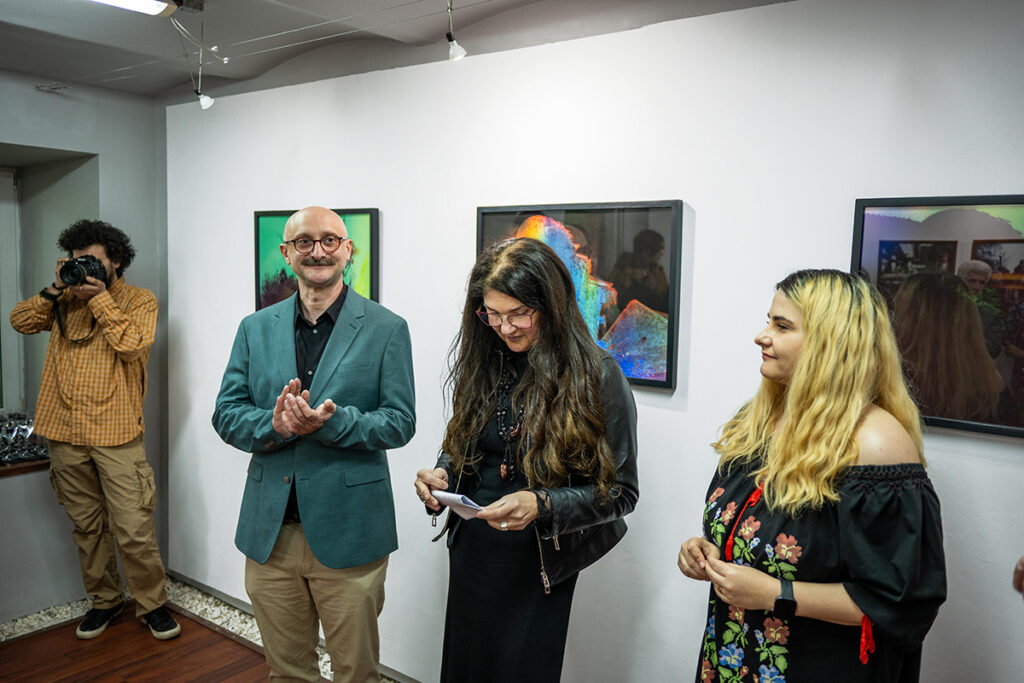 În imagine este artista Hermina Csata, alături de artiștii Mira Marincaș și Havadi-Nagy-David István. Un instantaneu de atmosferă de la vernisajul expoziției Q (curios, criptic, cataleptic) organizat de către Galeria Vault One & Korunk din Cluj-Napoca, în partneriat cu revista de fotografie Nophto, curator Teff Hășmășan.